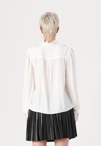 Bijela bluzica dugih rukava s ruffles na ovratniku i detaljima na ramenima. Uparen s crnom plisiranom suknjom. Glatka tkanina, ležeran dizajn.