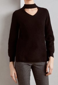 Czarny długi sweter z dzianiny z długim rękawem, dekoltem w serek i ozdobą typu choker, zestawiony z szarymi spodniami o jodełkowym wzorze.