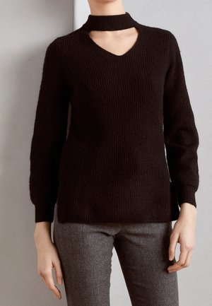 Zwarte, lange mouw, gebreide trui met een V-hals en choker-detail, gecombineerd met grijze broek met visgraatmotief.