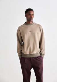 Man wearing beige crewneck sweatshirt and maroon trousers standing with hands in pockets against plain white background