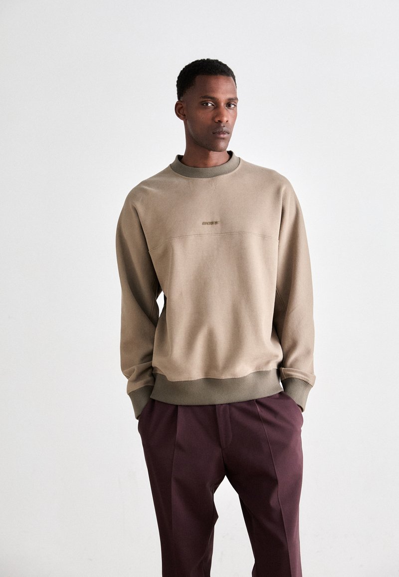 Man wearing beige crewneck sweatshirt and maroon trousers standing with hands in pockets against plain white background