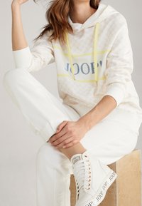 Hooded sweatshirt in light beige with a patterned design, yellow accents, and logo, paired with white joggers and high-top sneakers.