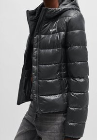 Svart puffjacka med glansig yta, utrustad med huva, vadderad design, dragkedja framtill och logotypdetalj på bröstet.
