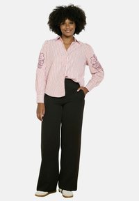 Pruhovaná ružová košeľa na gombíky s vyšívaným kvetinovým motívom na jednom rukáve, skombinovaná s čiernymi širokými nohavicami a bielou obuvou.