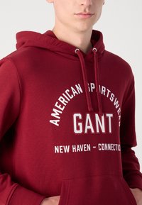 Bordó kapucnis pulóver, puha anyagból, fehér felirattal: "AMERICAN SPORTSWEAR GANT NEW HAVEN - CONNECTION." Elülső kenguruzseb.