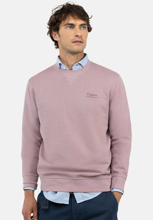 Hombre con una sudadera rosa claro sobre una camisa azul clara con botones y pantalones azul oscuro, mirando ligeramente hacia un lado.