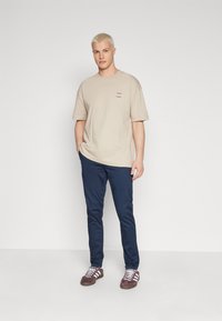 Beige bomull t-skjorte med korte ermer og liten tekstdetalj, kombinert med slim-fit marineblå bukser og brune joggesko med hvite striper.