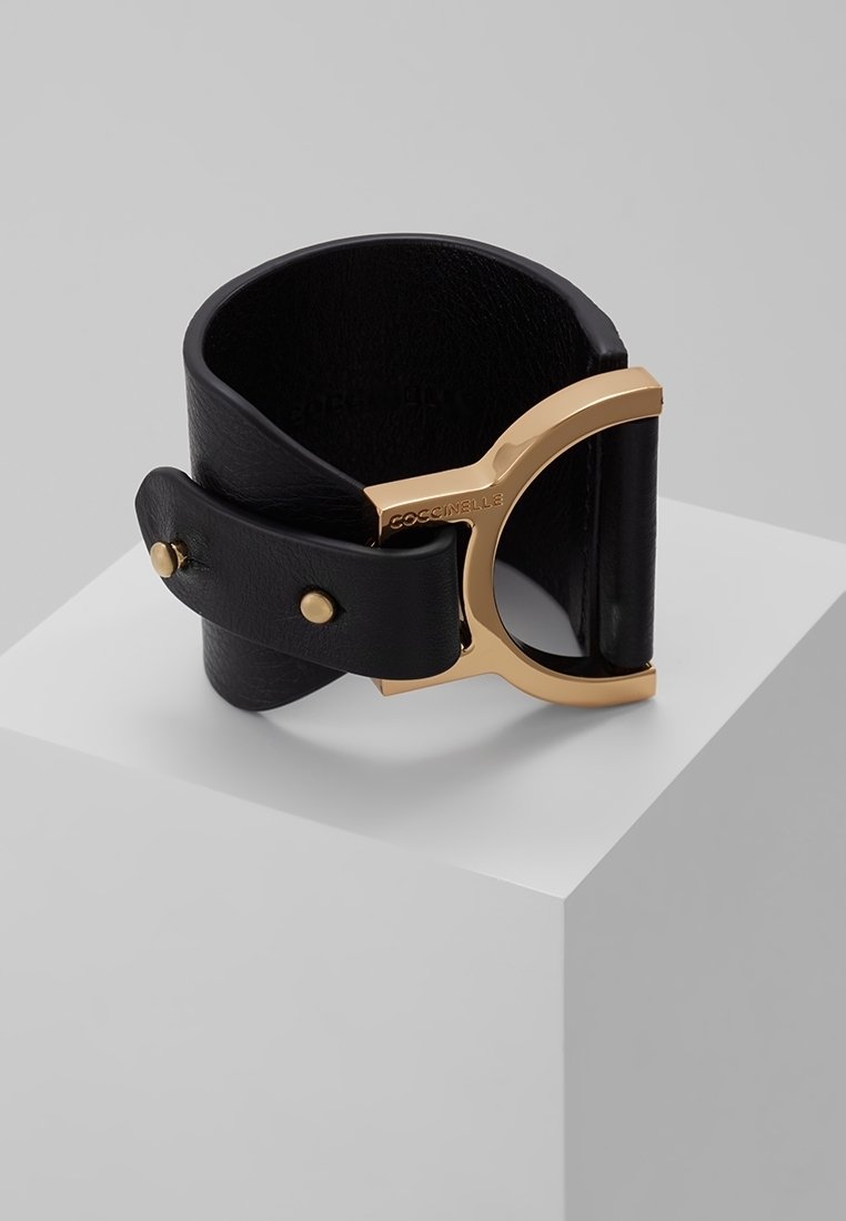 Bracelet manchette en cuir noir avec une large lanière et un grand fermoir doré gravé du nom de la marque, présenté sur un socle blanc.