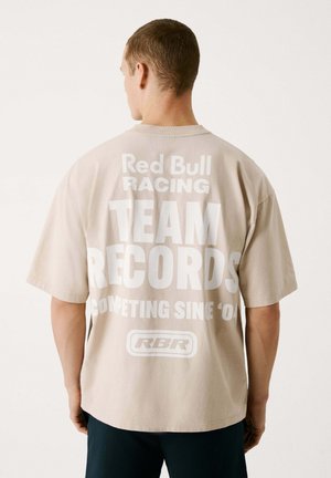 Beige übergroßes T-Shirt mit großem weißen Text auf der Rückseite: "Red Bull Racing TEAM RECORDS COMPETING SINCE '04", mit Logo darunter.