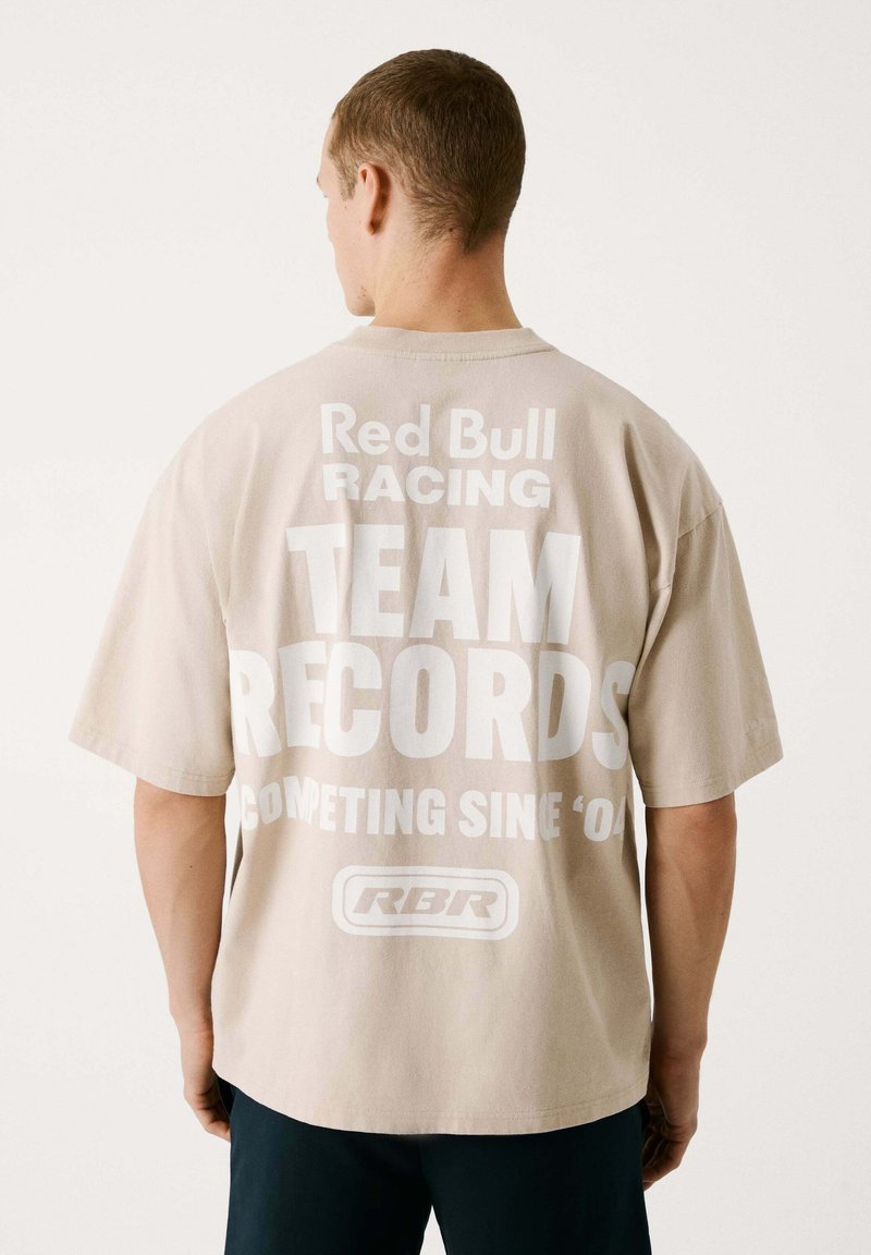 Beige übergroßes T-Shirt mit großem weißen Text auf der Rückseite: "Red Bull Racing TEAM RECORDS COMPETING SINCE '04", mit Logo darunter.