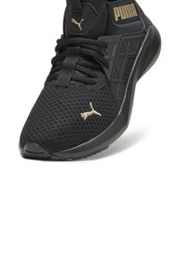 Chaussure de sport noire Puma en tissu mesh, fermeture lacet, avec logo Puma doré sur l’avant et la languette, inclinée pour montrer le côté et le devant.