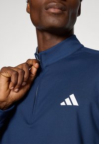 Námořnická modrá sportovní zipová mikina s vysokým stojáčkem a bílým logem adidas; hladká tkanina s přiléhavým designem, zobrazená částečně zapnutá.
