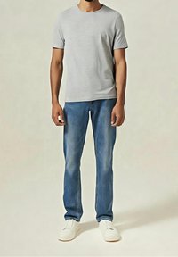 Homme portant un t-shirt gris clair à manches courtes, un jean bleu et des baskets blanches, debout devant un fond uni.