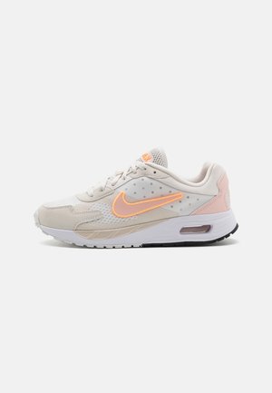 Vaalea beige Nike-tossu persikanvärisellä swooshilla, valkoinen välipohja näkyvällä ilmayksiköllä ja verkkopäällinen nauhoilla.