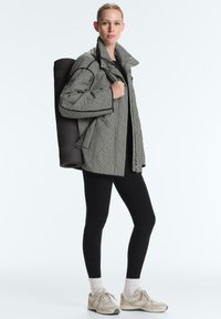 Femme debout portant des leggings noirs, des baskets beige, une veste à carreaux noire et blanche, portant un tapis de yoga noir roulé sur le dos.