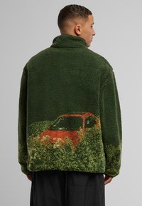 Grüne Fleecejacke mit einem Grafikmotiv eines orangefarbenen Autos vor einem grasbewachsenen Hintergrund, hohem Kragen und strukturierter, plüschiger Oberfläche.
