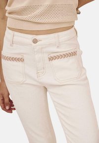 Jeans de mezclilla blanca con bolsillos traseros que presentan un detalle de bordado trenzado. Diseño de cintura alta con un cierre de botón clásico en la parte frontal.