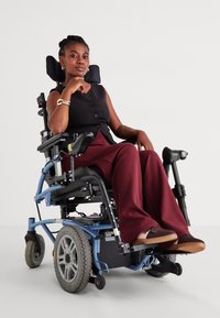 Fauteuil roulant électrique avec un cadre métallique bleu et de grandes roues. L'utilisateur porte un haut sans manches noir et un pantalon bordeaux, assis avec confiance.
