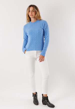 Donna con capelli biondi mossi, che indossa un maglione blu texturizzato, pantaloni bianchi e stivaletti neri alla caviglia, in piedi con una mano in tasca.