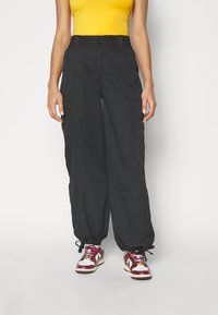 Svarta cargobyxor med elastisk midja, flera sidofickor och justerbara muddar. Bärs med röda och vita sneakers och en gul topp.