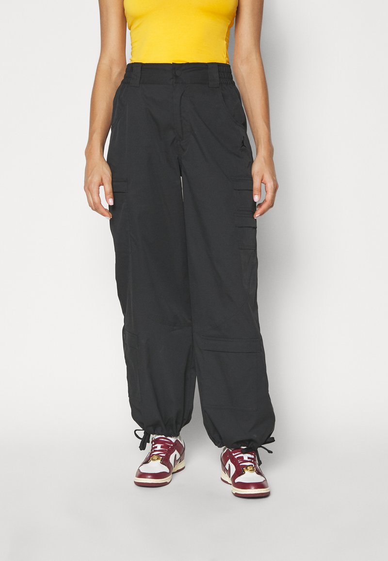 Svarta cargobyxor med elastisk midja, flera sidofickor och justerbara muddar. Bärs med röda och vita sneakers och en gul topp.