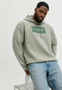 Zielona bawełniana bluza z kapturem, z sznurkiem i logo z przodu, w połączeniu z jasnoniebieskimi dżinsami. Model opiera się o metalowe krzesło.