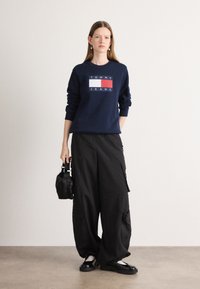 Felpa della marina con il logo "TOMMY JEANS" in bianco e rosso. Pantaloni cargo larghi neri. Modella che tiene una piccola borsa nera, indossando scarpe nere.