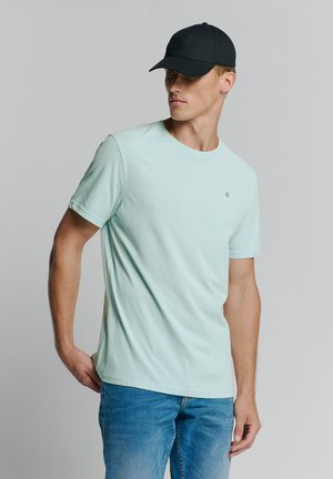Licht mintgroen T-shirt met korte mouwen en een klassieke ronde hals, gecombineerd met blauwe jeans en een zwarte baseballpet. Subtiele logo-detail op het T-shirt.