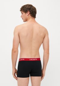 Des boxers noirs avec une ceinture élastique rouge portant le logo "LACOSTE". Le tissu semble lisse avec un ajustement serré, mettant en valeur la silhouette du corps.