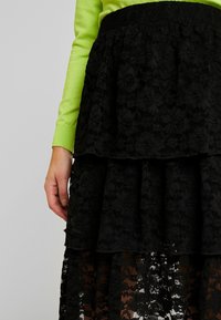 Jupe en dentelle noire avec un design superposé, présentant un motif floral et un tissu texturé, associée à un haut à manches longues vert vif.