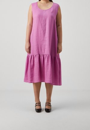 Mujer con vestido rosa sin mangas hasta la rodilla con dobladillo fruncido y zapatos Mary Jane negros de doble correa, posando frente a un fondo gris liso.