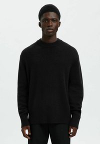 Joven de piel oscura que lleva un suéter negro sencillo y pantalones negros, de pie contra un fondo claro y liso.
