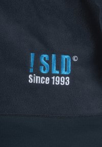 Tejido de forro polar oscuro con texto bordado en blanco y azul claro: "! SLD © Desde 1993" centrado. Textura suave, sin patrones.
