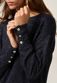 Granatowy sweter z metalicznymi drobinkami, z okrągłym dekoltem i mankietami zapinanymi na guziki ozdobione jasnoniebieskimi i przezroczystymi guzikami.