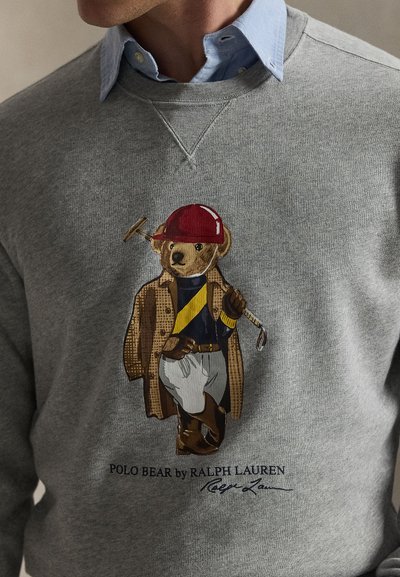 Γκρι φούτερ με κεντημένο αρκούδο που φοράει καρό παλτό και κόκκινο καπέλο, με μπλε πουκάμισο από κάτω και μπαστούνι γκολφ στο πόδι.
