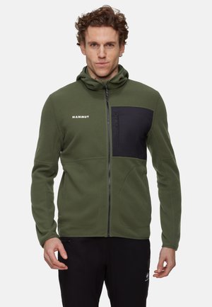 Mężczyzna w zielonej polarowej kurtce na zamek z czarną kieszenią na piersi i logo "Mammut" na lewej piersi, stoi na jednolitym tle.