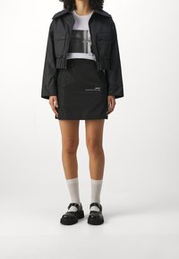 Černá krátká bunda s kapsami na knoflíky, vrstvená přes bílá tričko s potiskem. Černá mini sukně s nápisem, doplněná černými botami na přezku a ponožkami.