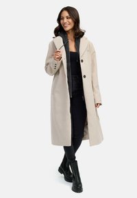 Beige lange jas met grote revers, enkele knoopsluiting, zijzakken, gelaagd over een donkere hoodie en zwarte jeans, gecombineerd met zwarte laarzen.
