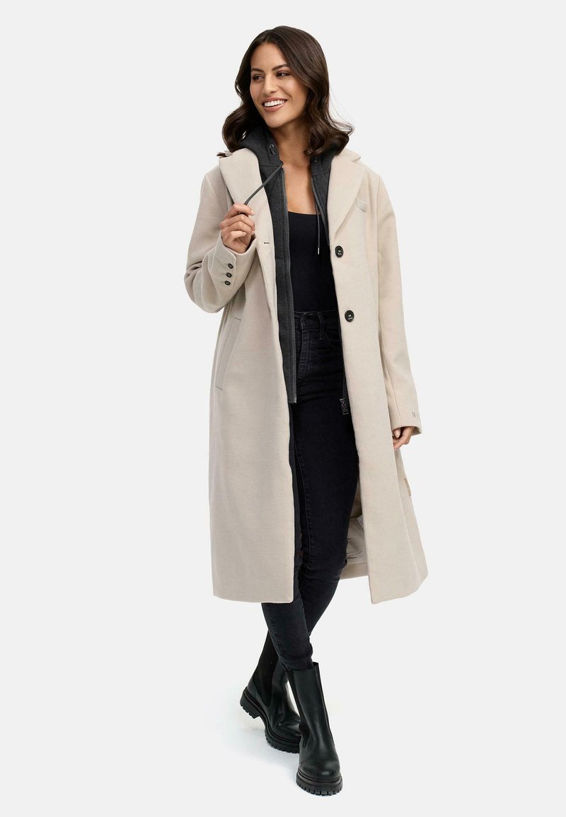 Beige lange jas met grote revers, enkele knoopsluiting, zijzakken, gelaagd over een donkere hoodie en zwarte jeans, gecombineerd met zwarte laarzen.