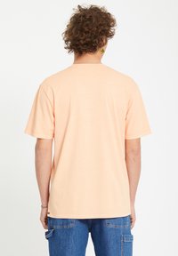 Lys ferskenfarvet kortærmet t-shirt lavet af blødt stof, med rund hals og en afslappet pasform, set fra ryggen. Et par jeans bæres nedenfor.