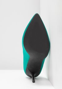 Tyrkysová vysoká bota na podpatku s špičatou špičkou a štíhlým stiletto podpatkem. Podešev je černá s texturovaným povrchem pro lepší přilnavost.