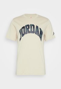 Beżowa bawełniana koszulka z dużym, łukowatym logo "JORDAN" w ciemnozielonym i czarnym kratkowanym wzorze, z małym czarnym logo w sylwetce na rękawie.