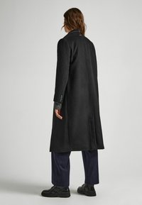 Abrigo negro largo de corte recto, cuello con muesca y tejido texturizado. Combinado con pantalones azul marino de rayas diplomáticas y zapatos negros con cordones.