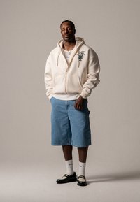 Tejfehér zipzáras kapucnis pulcsi zöld hímzéssel, farmer rövidnadrág, fekete és fehér cipők, fehér zoknik és ezüst nyaklánc. Minimalista dizájn.