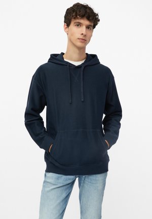 Junger Mann mit lockigem Haar, der einen navyblauen Hoodie und hellblaue Jeans trägt, steht mit den Händen in den Taschen des Hoodies vor einem schlichten Hintergrund.