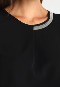Prenda negra con escote redondo que presenta un acento plateado en el cuello. La textura parece suave y ligeramente drapeada.