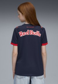 Maillot de sport navy avec des accents rouges, présentant "Red Bulls" en lettres blanches en gras au dos et le texte "DES IS SOIZBURG" au-dessus.