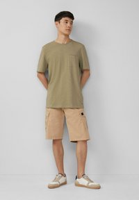 Olijfgroen t-shirt met korte mouwen en een borstzak, gecombineerd met lichtbeige cargoshorts met zijzakken en een zwart vierkant label. Witte sneakers.