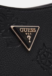 Surface noire texturée avec une plaque triangulaire noire Guess USA bordée d’or, avec un point d’interrogation doré sous le texte.