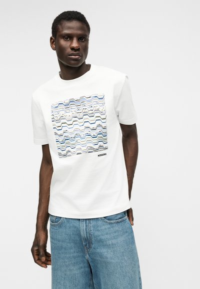 Άνδρας που φοράει λευκό κοντομάνικο T-shirt με μπλε και μπεζ ζιγκ-ζαγκ σχέδιο και το κείμενο "MISSONI", συνδυασμένο με ανοιχτού μπλε τζιν, στέκεται μπροστά σε λευκό φόντο.